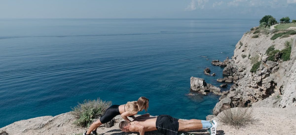 A couple performs a partner workout on a scenic cliffside, overlooking the ocean.